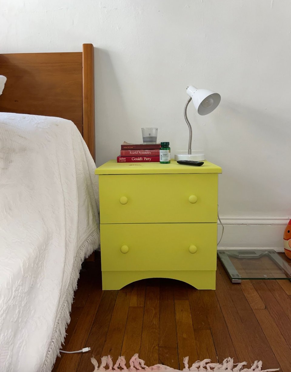 A bright yellow-green side table with a lamp and stack of books, standing beside a bed.