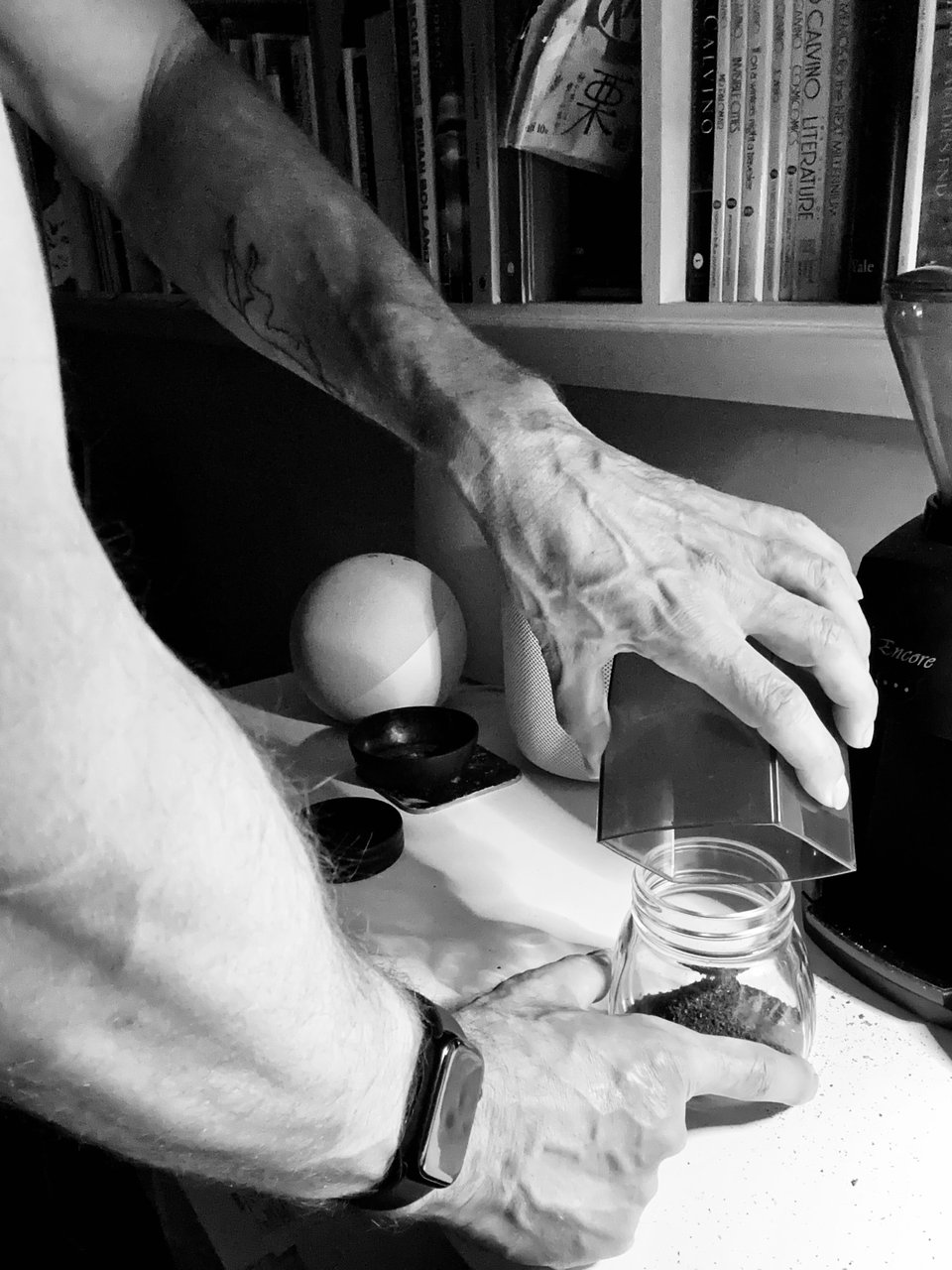 b/w photo of white man's hands/forearms as he pours ground coffee into a container, in front of bookshelves