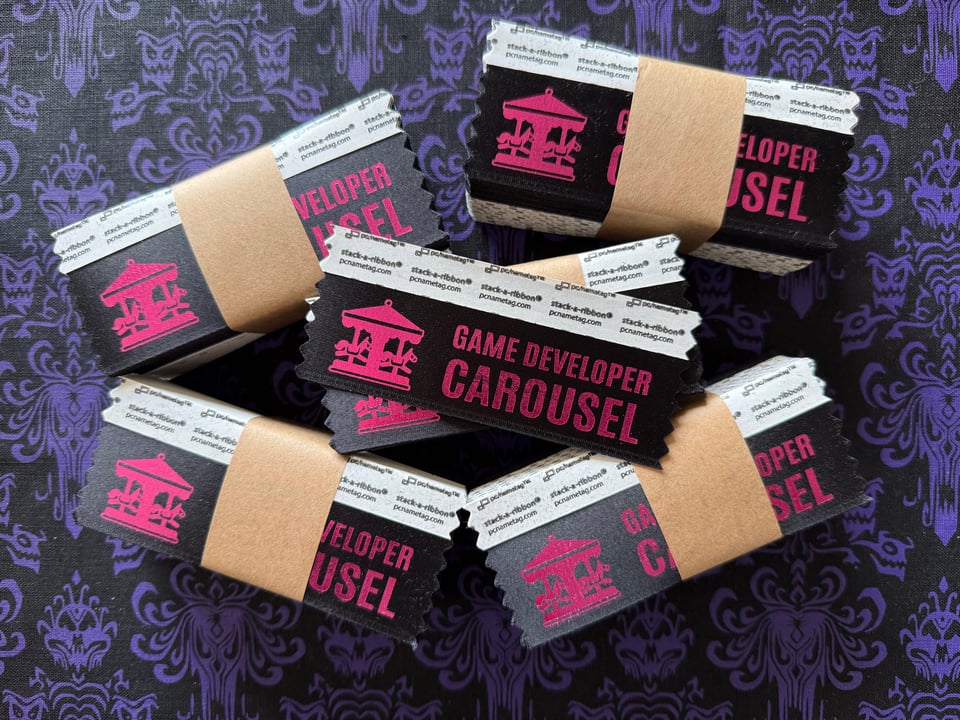 Five stacks of badge ribbons with one single ribbon on top of the stack. Each ribbon has the words Game Developer Carousel and the logo in pink on a black ribbon.