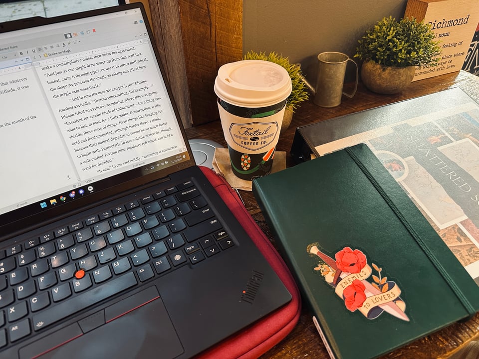 Cass's laptop on a coffee shop table next to a branded cup, a dark green bullet journal with an "Enemies to Lovers" sticker on it, and a binder with a manuscript-themed collage on the front.