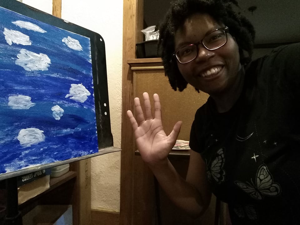 An image of a young Black woman with a bright smile, waving hello. She wears glasses and a black shirt with a butterfly pattern. Beside her, there is a blue painting of clouds in a sky.