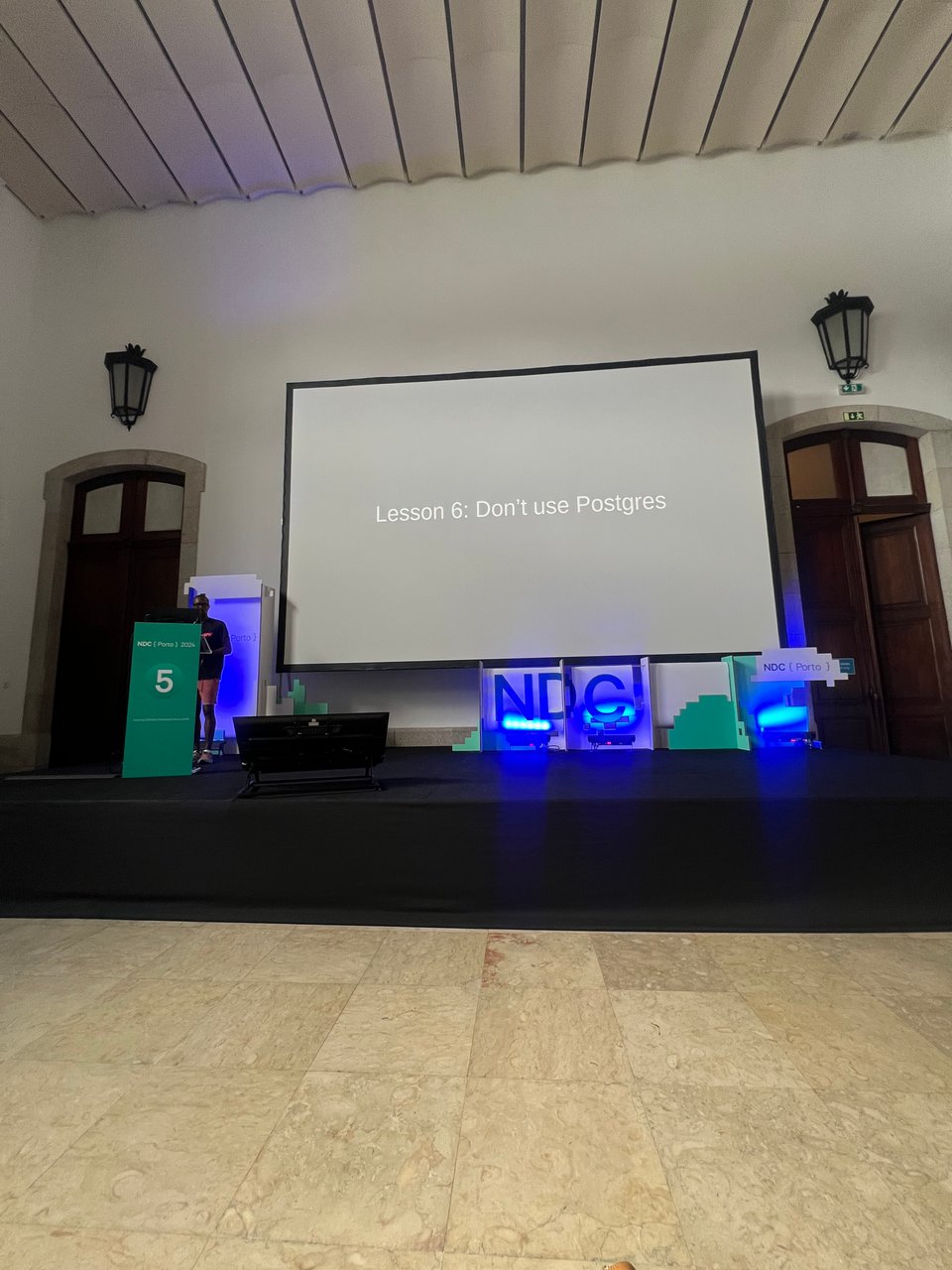 My cofounder Rob is presenting at a tech conference, standing next to a slide that reads "Lesson 6: Don’t use Postgres." The room is spacious with traditional architectural details.