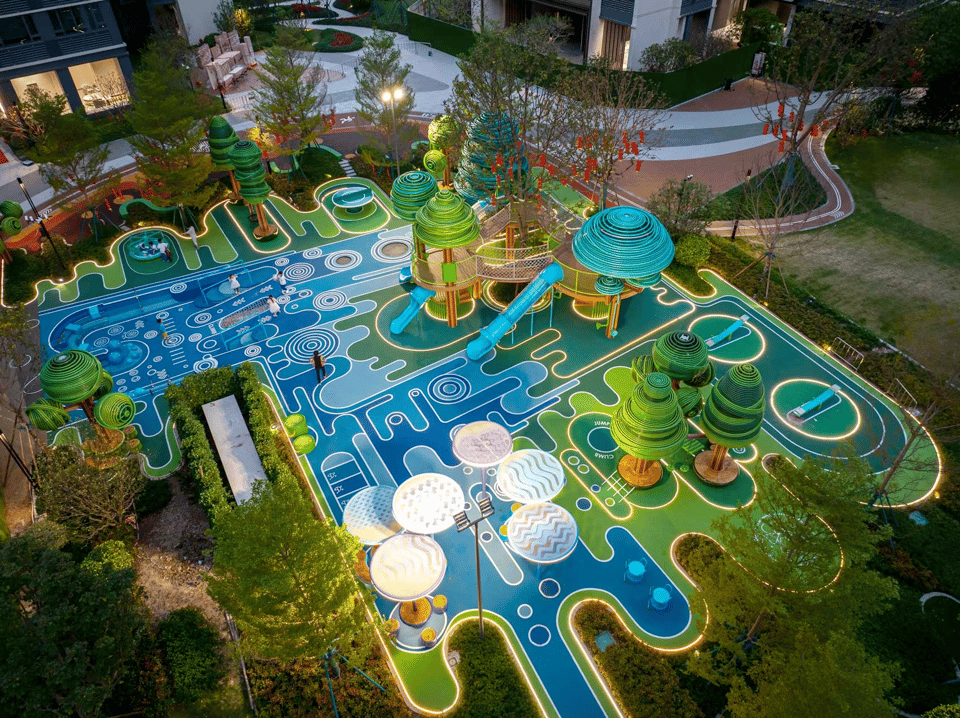 A futuristic and vibrant playground photographed from above at twilight, featuring an elaborate design with glowing outlines and colorful surfaces. The ground is painted in shades of blue and green with intricate patterns resembling circuit boards or abstract mazes. Tree-like structures with spiral canopies rise from the surface, some connected by bridges and slides. Circular seating areas, climbing structures, and swings are integrated into the landscape. The entire area is illuminated with embedded lights that trace the layout, creating a visually striking and imaginative environment. Surrounding the playground are landscaped paths, greenery, and nearby residential buildings.