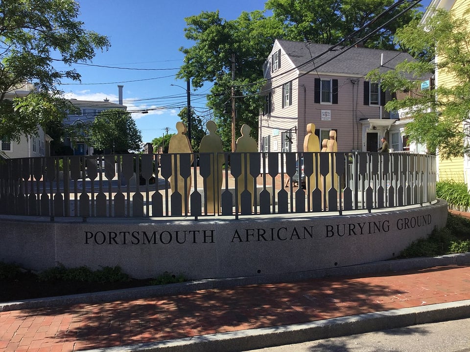 A sunny landscape photograph of a New England city park. In the foreground, the words PORTSMOUTH AFRICAN BURYING GROUND are carved in granite across a large platform, on which stand a stylized metal fence and the statues of six silhouetted figures, each representing an African person buried on the site.