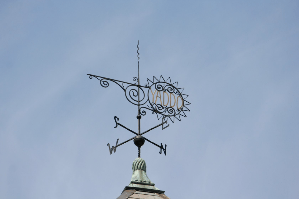 An ornate iron weathervane with the word Yaddo in the center against a light blue sky.