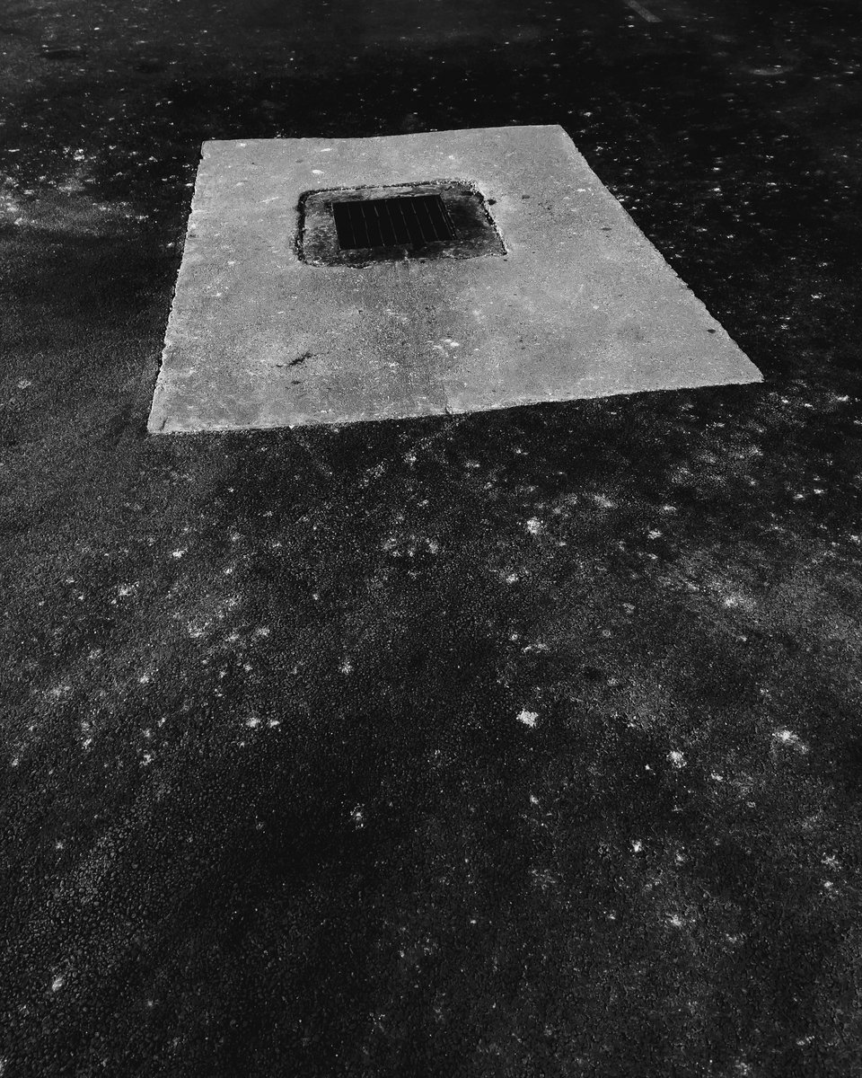 a square catch basin in a rectangle of concrete appears to float in a starry sky illusion created by salt stains on asphalt