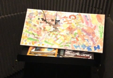 Picture of a music stand with a canvas on it. The canvas is painted with many colors, creating some kind of abstract art. There are two small microphones stuck to it to record the sounds of painting brushes.