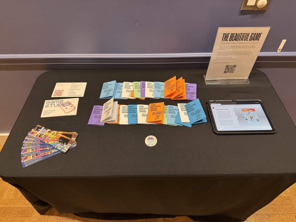 A display table covered with a black cloth holds rows of colorful "Old Games Zine 2026" booklets, fanned-out illustrated cards, small printed info cards, a round button, a tablet showing a webpage, and an acrylic sign reading "The Beautiful Game: A Playable History of Soccer Video Games" against a purple wall.