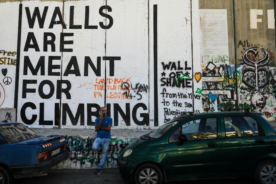 A West Bank apartheid wall covered in graffiti on the Palestinian controlled side. Some of the graffiti says "Walls are meant for climbing" "Land back" "Wall of Shame" "From the river to the sea Palestine will be free" and drawings and artwork.