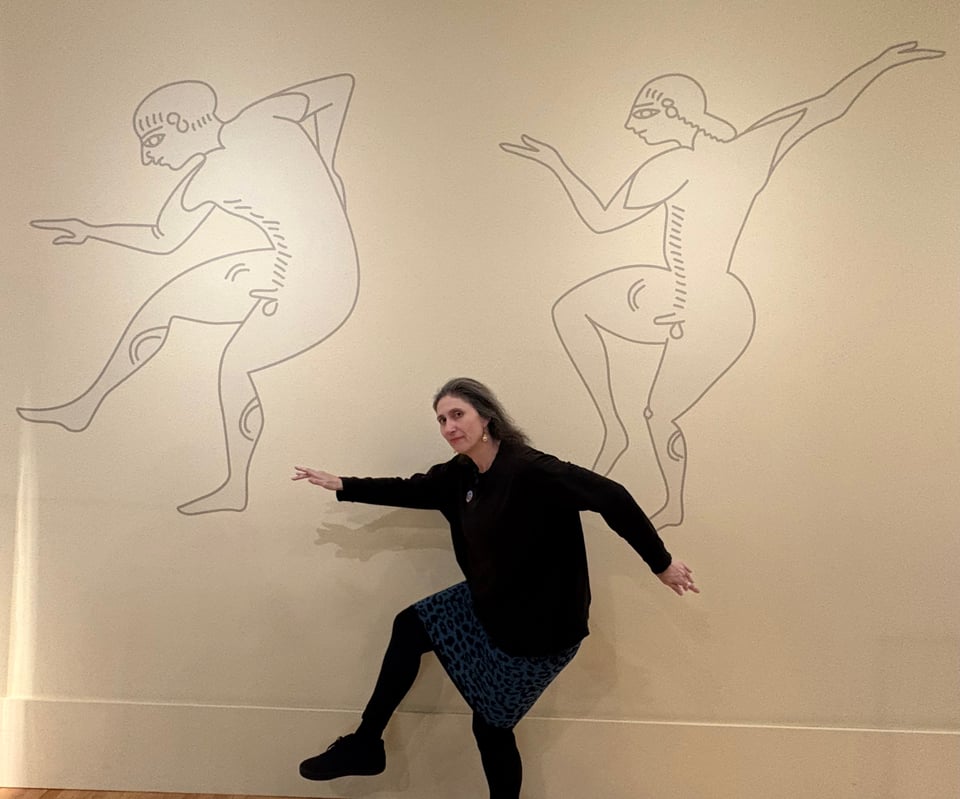 Alt text: A person poses in front of a wall with Etruscan inspired art on it.