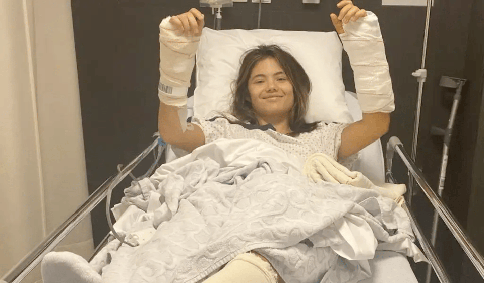 Emma Raducanu in a hospital bed, raising up both arms to show off casts around her wrists.