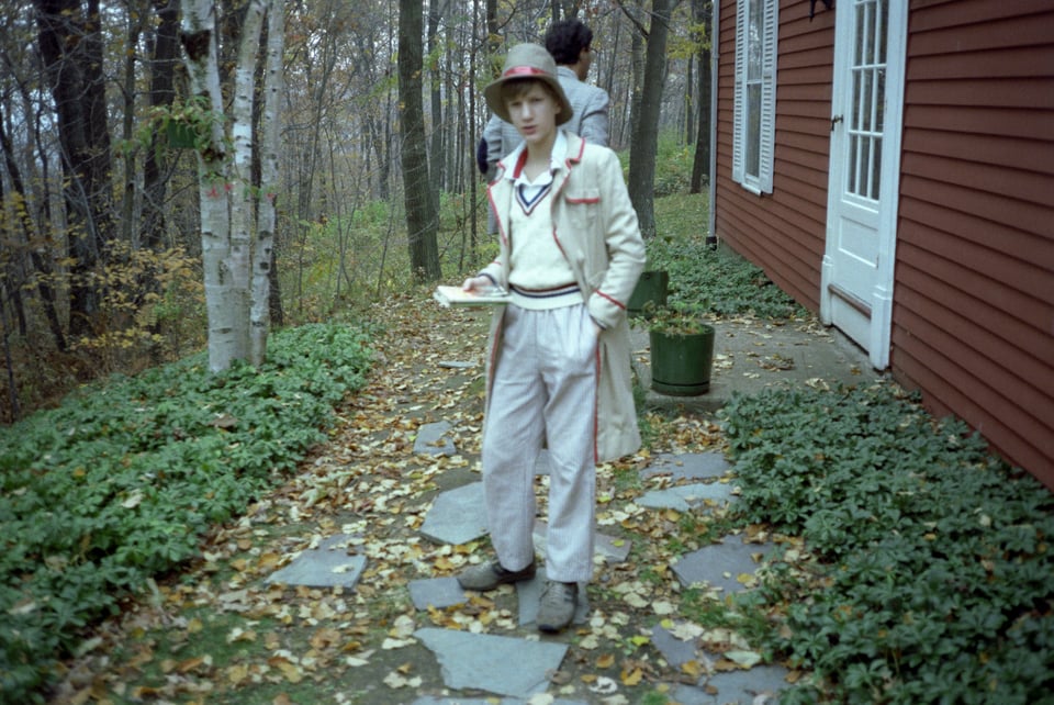 Charlie Jane as a young person, dressed as the Fifth Doctor from Doctor Who, standing in front of a red house on a path.