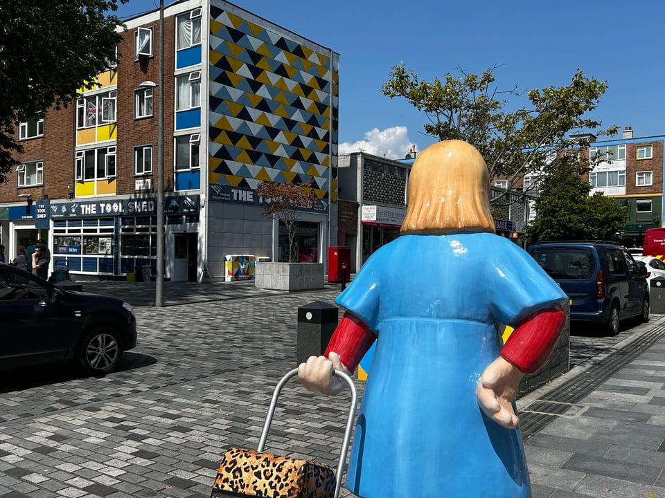A large fibre-glass model based on an Beryl Cook painting is pulling her leopard print shopper along a street. cross the road is a 1960s set of flats with a blank face decorated with yellow, blue, black and white triangles. The blue of the building matches the blue of the sculpture's dress.