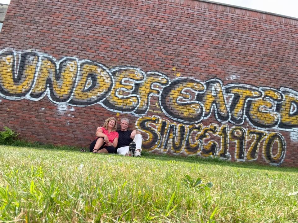 A white woman and man sitting on grass in front of a brick wall that has the graffiti "UNDEFEATED SINCE 1970"