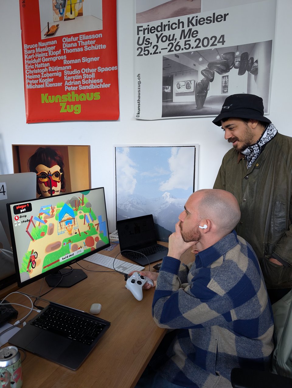 Two people - one seated, one standing - play a colourful game where a character is trying to build a shed