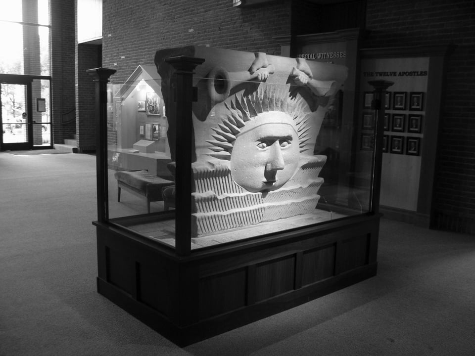 Black and white photo of a large piece of stone carved with a sun face and hands holding trumpets, in a display case in the Smithsonian.