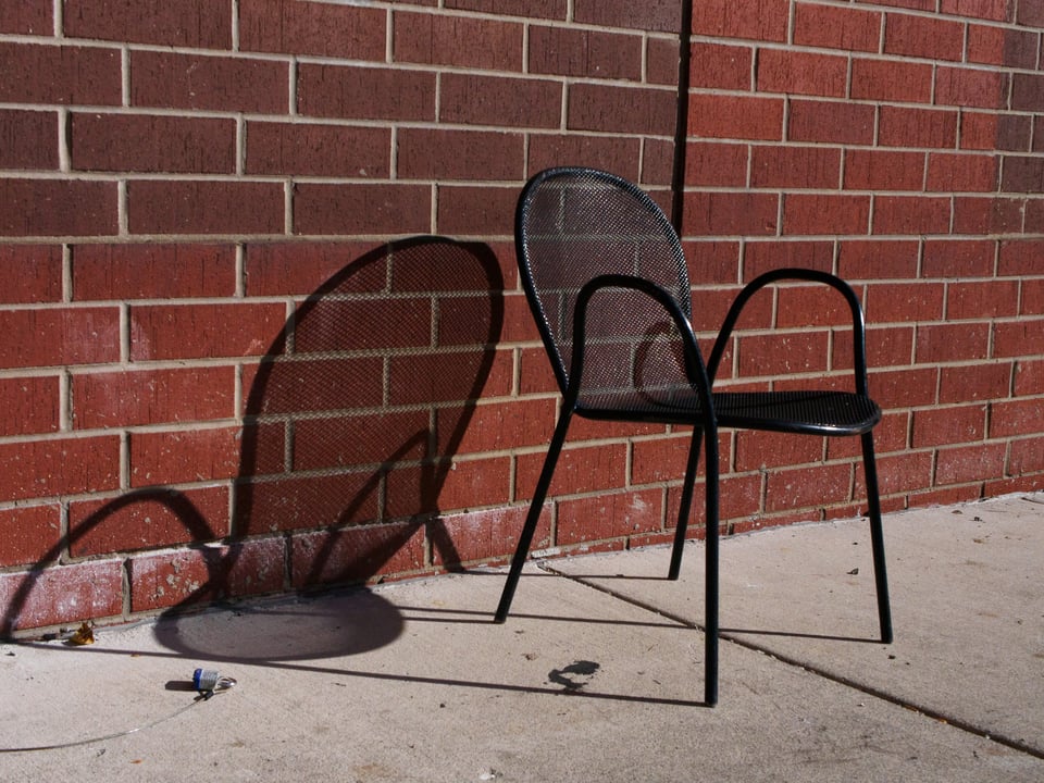 A black metal chair against a red brick wall. The chair is casting a very distinct shadow on the wall as well.