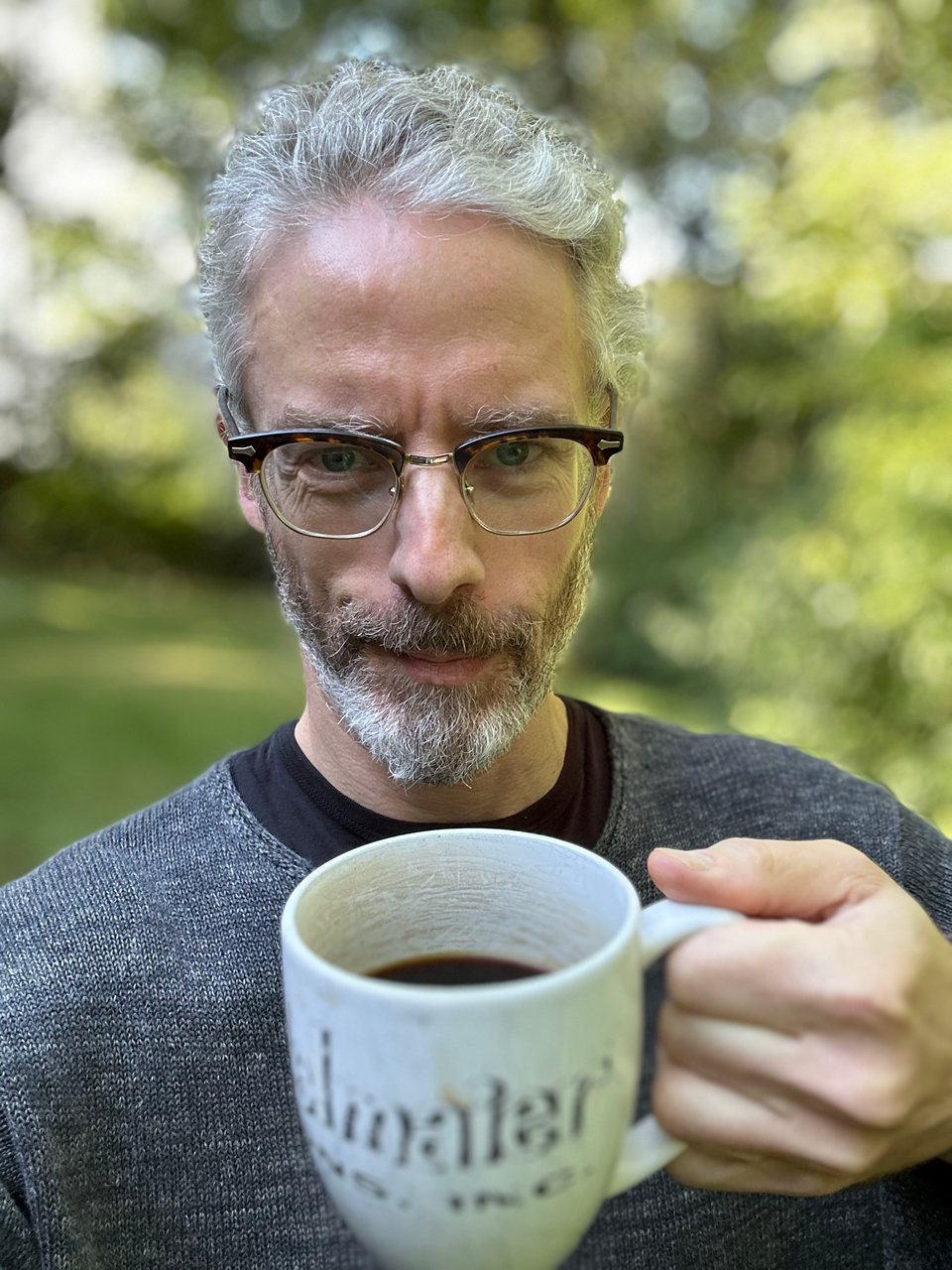 Photo of white man with beard and horn-rimmed glasses holding a big coffee mug up to the camera