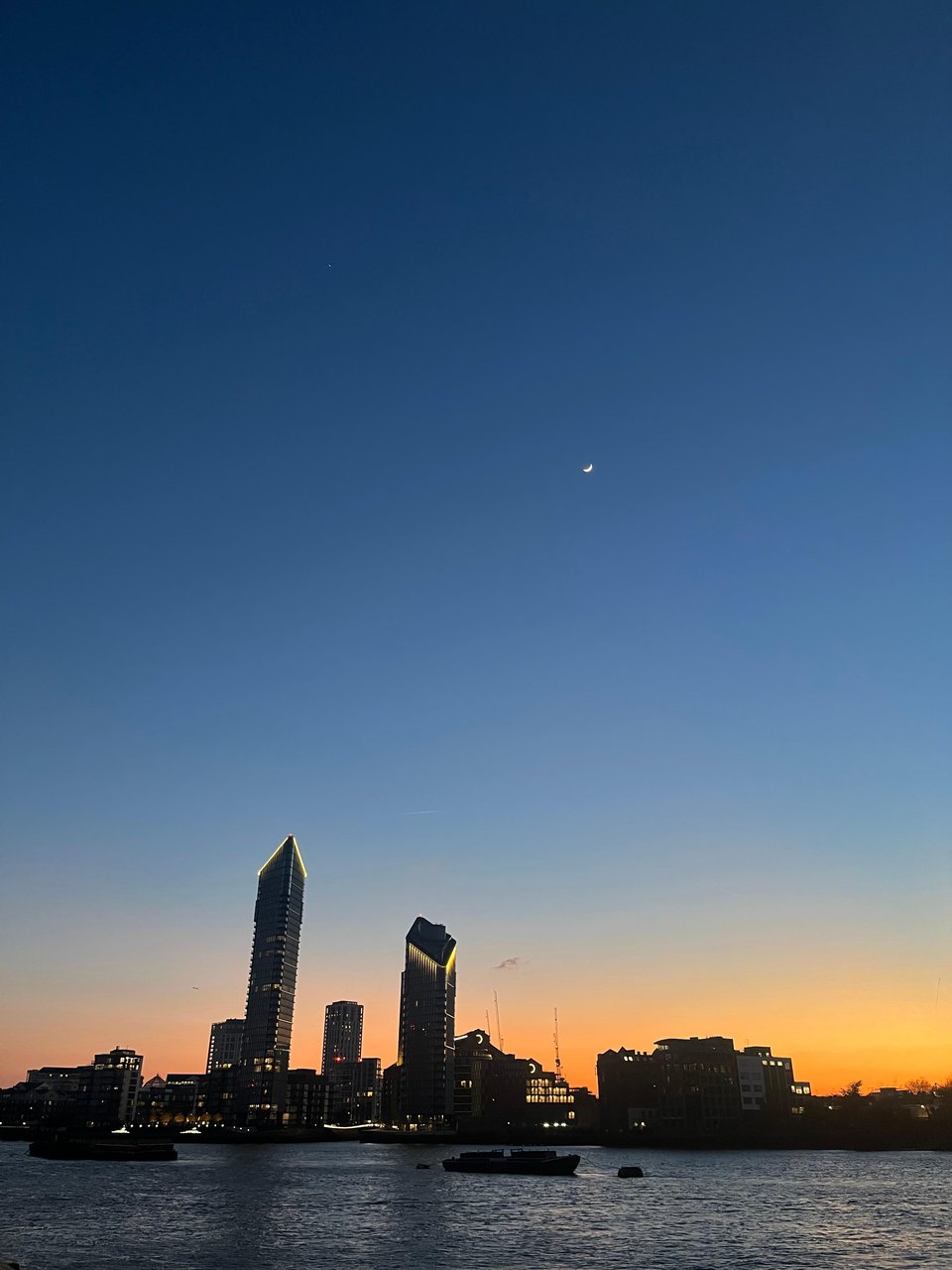 A photo of sunset above the Thames. At the bottom of the photo is a dark Thames and a silhouette of the London skyline (without any recognisable landmarks), with an orange glow behind it, which fades to a dark midnight blue as you move up the frame.