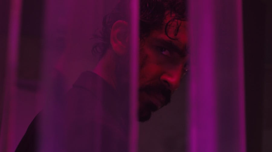 Dev Patel in a screenshot from Monkey Man, peering through sheer purple screens of some sort. He is wearing a black suit, his face looks battered, and he appears to be angry and getting ready to for revenge.