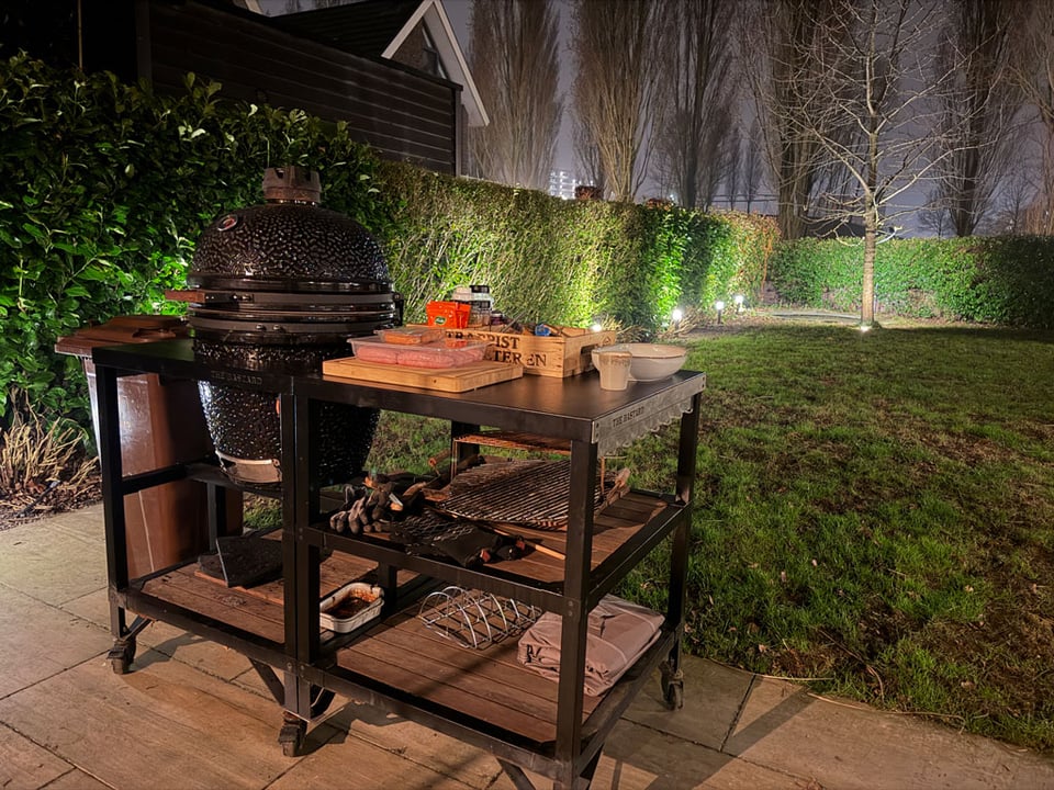 A nighttime scene - bbq on a moveable tray with a grass garden and green hedge all around behind it.