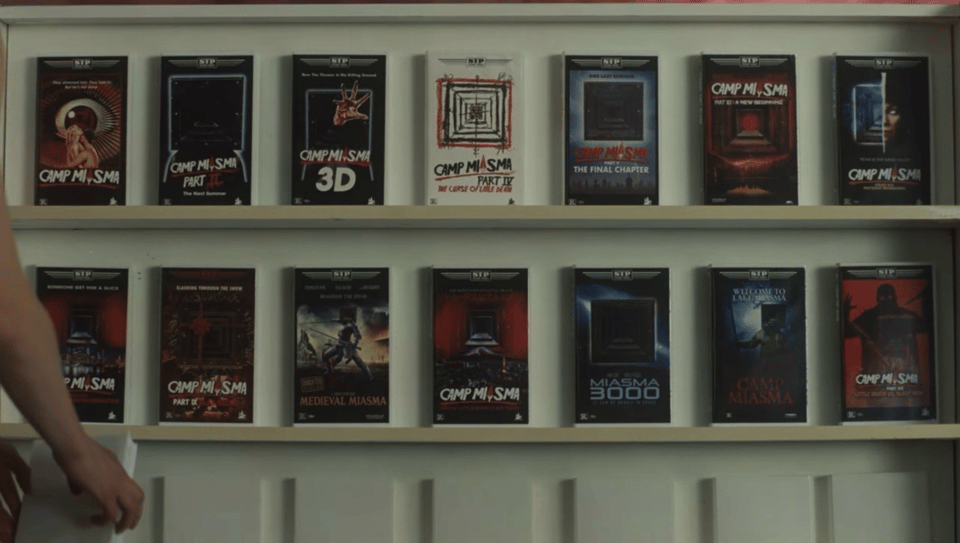 A shot from "Teenage Sex and Death at Camp Miasma" showing a shallow white shelf stocked with VHS tapes from the Camp Miasma series.