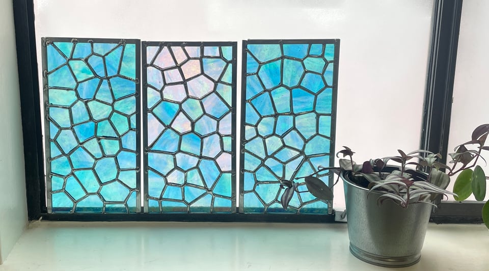 Three rectangular stained glass panels are sitting on a window ledge, backlit and glowing teal blue. The pieces are cut and assembled as an irregular cellular (Voronoi) pattern. A small houseplant next to the panels suggests scale.