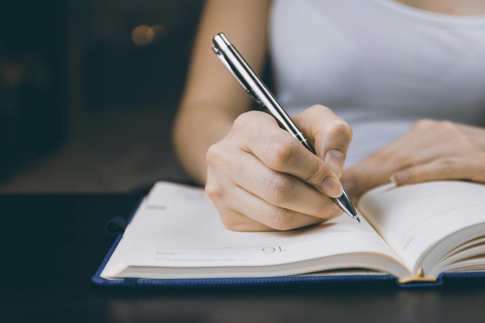 Stock photo of a person writing in an open notebook. (Face not shown)