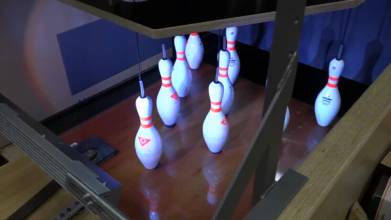 An animated gif showing a bowling ball striking 10 white bowling pins. The pins are hanging from blacks strings. When the ball hits the pins, they all scatter and lie on the ground for a second; then the strings pull on them and the pins all rise up into the air where they fit into holes in a piece of wood that hold them steady, before they drop down again