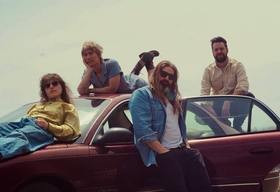 Four people lie atop, or stand next to, a red four-door sedan with the right rear door open on a hazy day.
