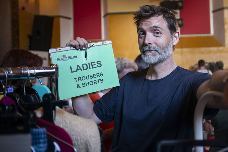 Patrick Grant holding an A4 sign advertising ladies' trousers and shorts, alongside the Action on Climate Emergency Settle and area logo