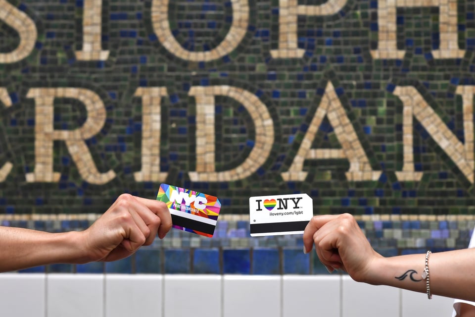 Arms of two people each holding commemorative rainbow-flag themed MetroCards in front of the tile mosaic at Christopher Street-Sheridan Square subway station.
