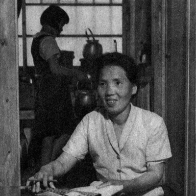 a photograph of a woman sitting at a table writing in a book, in the background someone can be seen in the kitchen