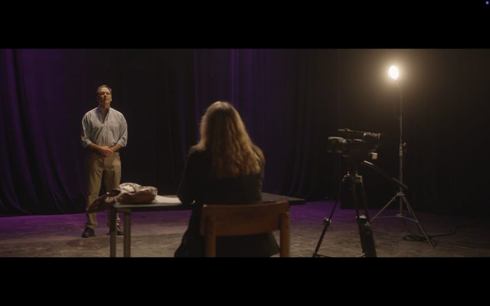 e of film. A woman sits at a desk. A man stands nervously in front of her as a camera stares and a light glares