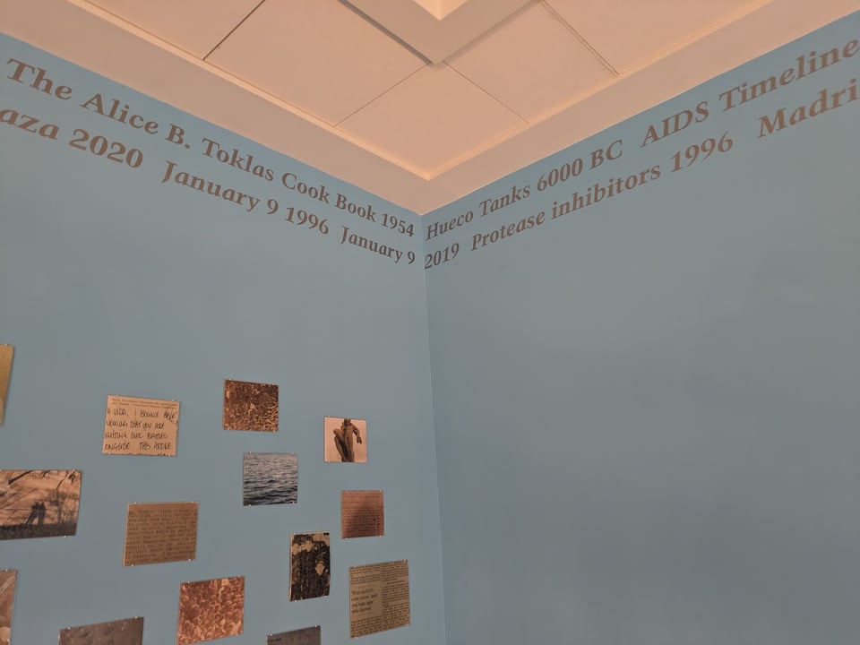 Light blue gallery walls feature words and dates in silver paint along the ceiling line, and photographs scattered on the wall.