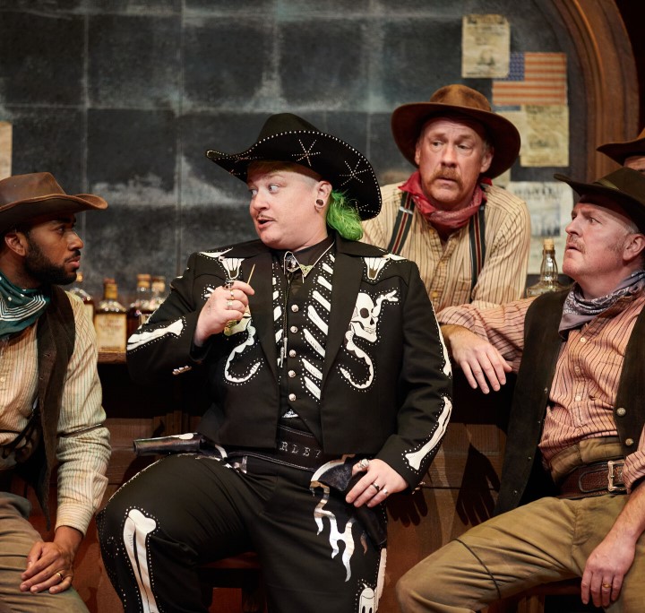 A broad cowboy with a green mullet and a black and white skull covered outfit surrounded by attentive cowboys in more earth toned looks