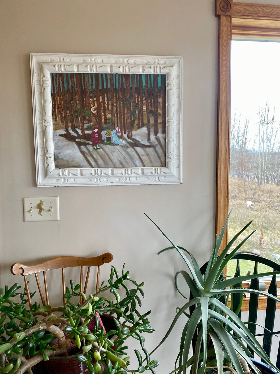 an art of kcf painting hanging in their home with two indoor plants below the painting and a sliver of a window showing an outdoor scene with two goats grazing on a hill