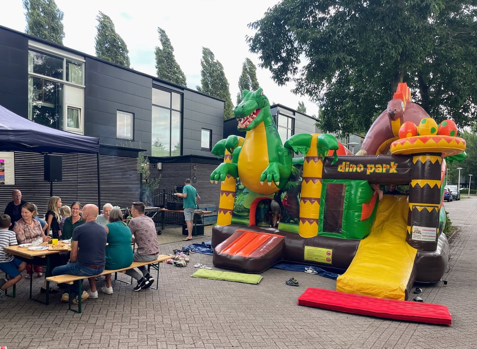 A street is blocked off with a large bouncy castle and people sitting at tables and benches enjoying a BBQ.