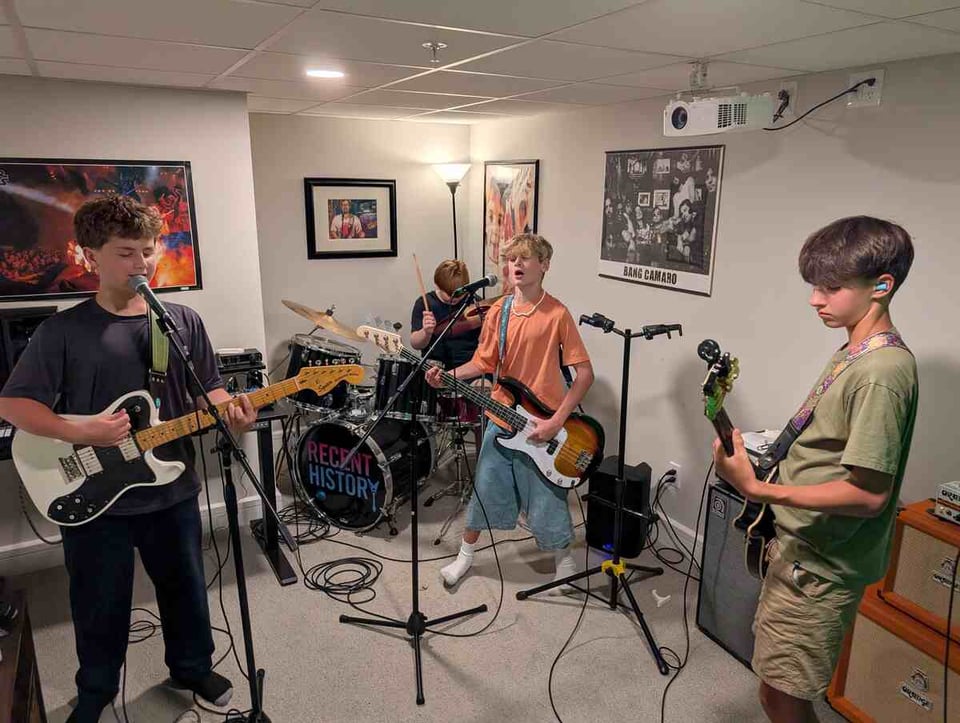 Four adolescent boys play instruments in a basement rec room. The boy at left plays electric guitar. To his left at the back, a boy plays drums, with a bassist in front of the kit. On the right side of the frame, another boy plays guitar.