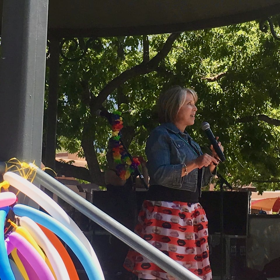 Governor Michelle Lujan Grisham at the microphone on the Santa Fe Plaza bandstand, celebrating Santa Fe Pride. She's wearing her denim jacket, a red and white checked skirt with lips on it and red boots.
