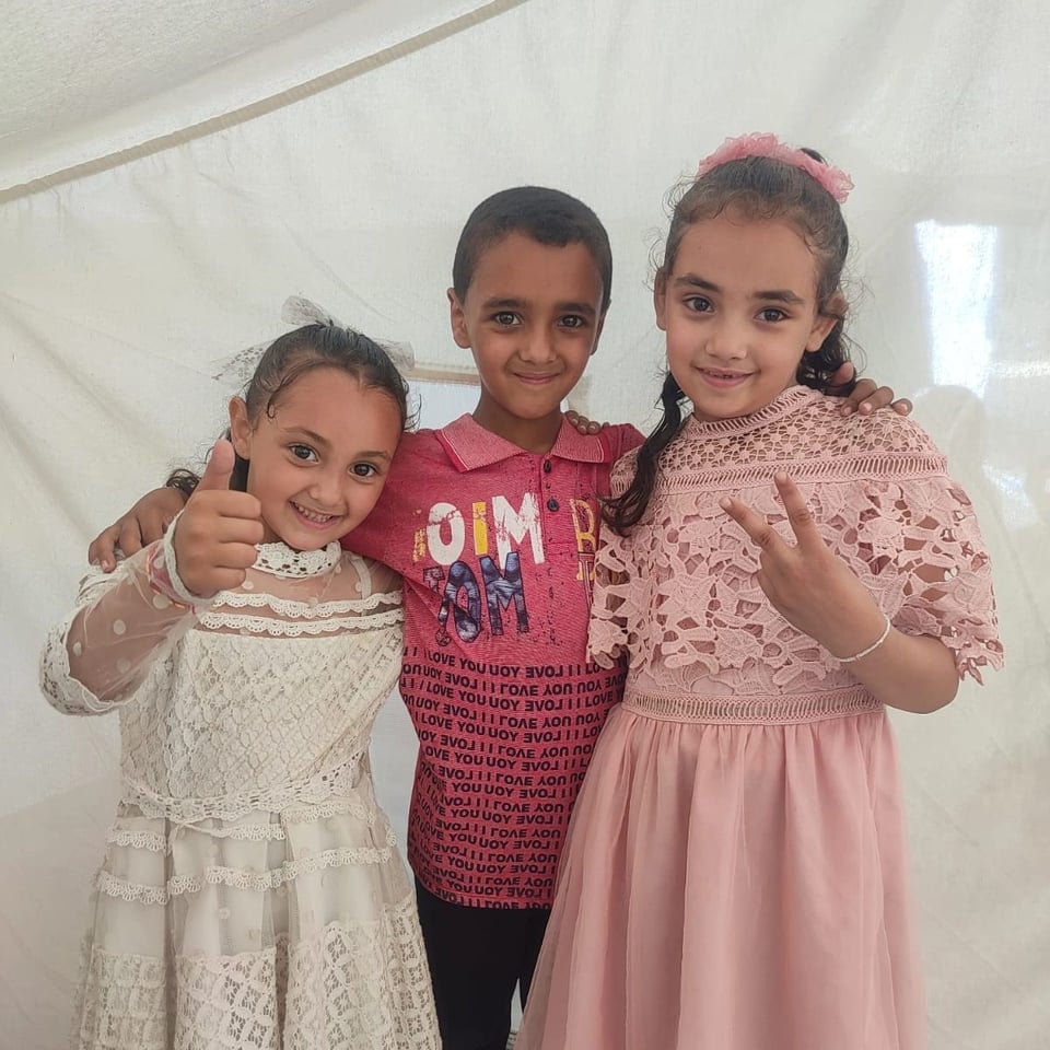 Three smiling children stand together in front of a light-colored backdrop. The boy in the center wears a red shirt with text, while the two girls beside him wear lace dresses—one in white giving a thumbs-up and the other in pink making a peace sign.