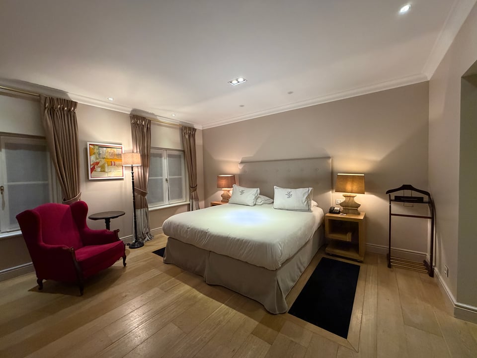 Hotel bedroom with a double bed, nightstands, a nice red chair and windows with curtains pulled back.