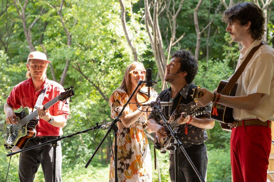 Four people perform music together in front of a background of trees: from left, a man with a resonator guitar, a woman playing fiddle, a man playing mandolin and a man playing guitar.