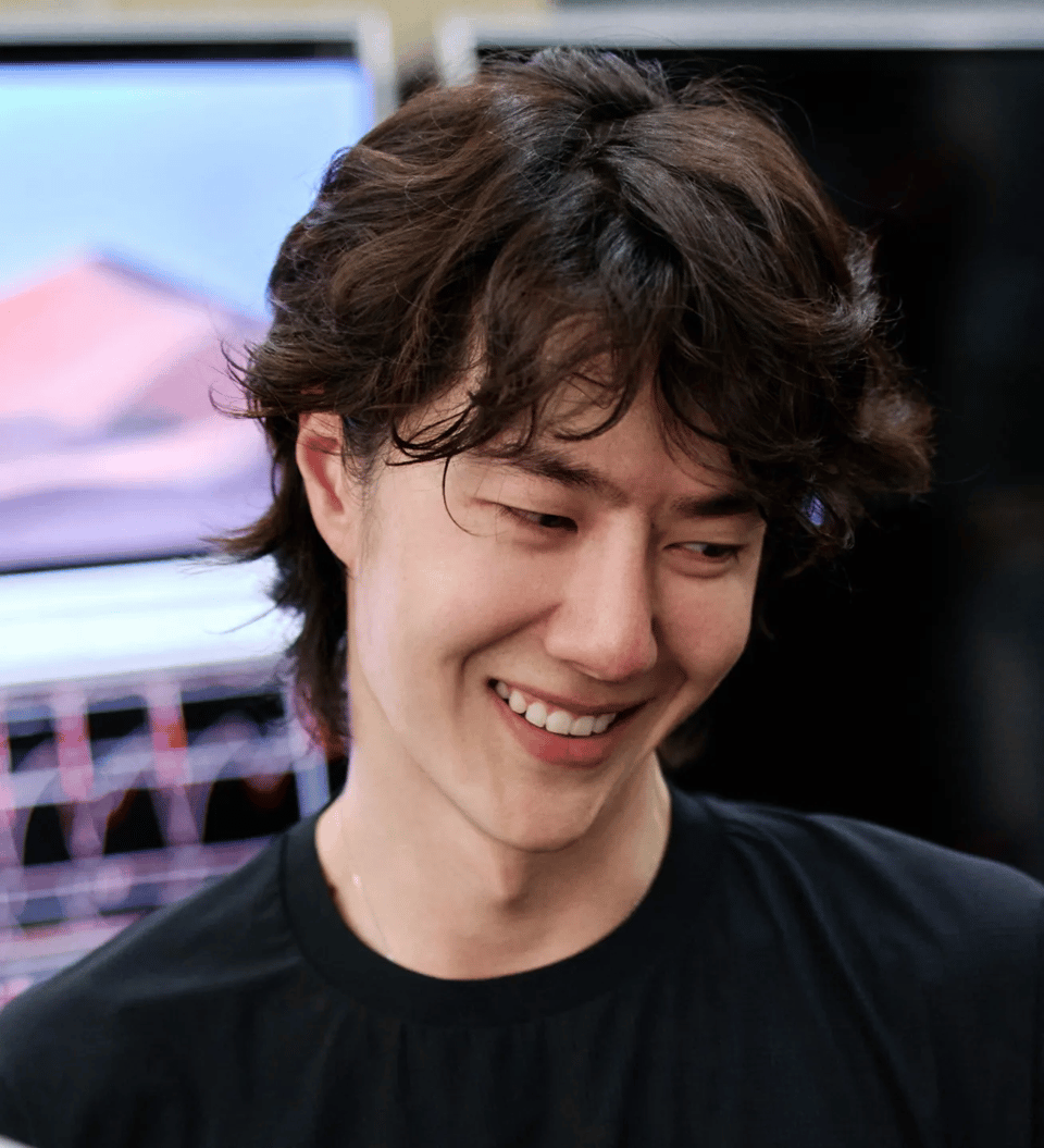 Photo of Wang Yibo in a black t-shirt with curly long-ish hair, looking down and grinning from ear to ear.