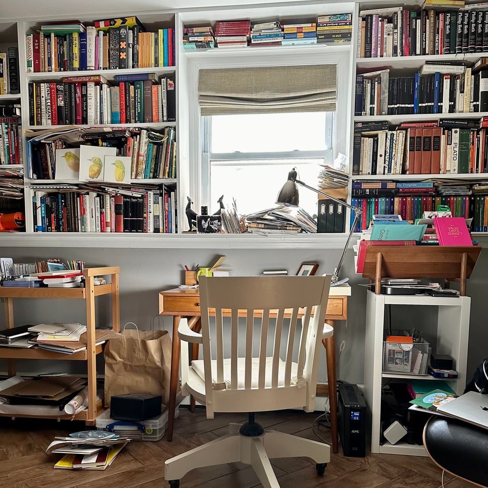 Photo of a library/office, centered on a white wooden chair in front of a small desk. There are shelves filled with books, an art-cart, and a window in the background
