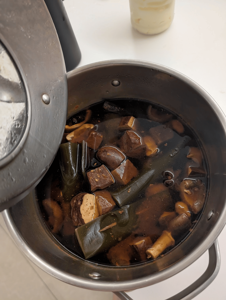A pot of soup - the broth is very dark brown and it contains chunks of tofu, kombu (on toothpick skewers) and shiitake mushrooms.