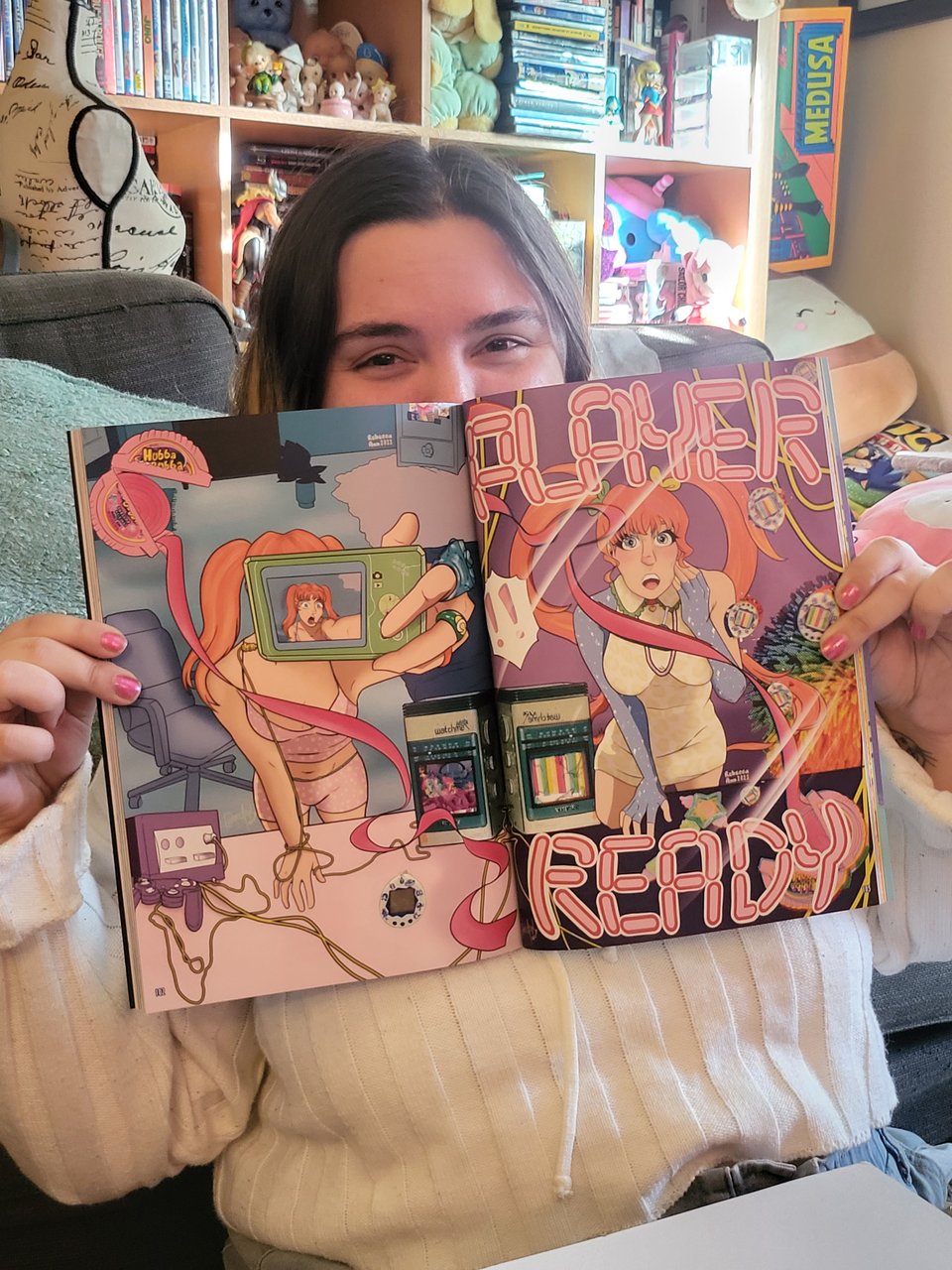 A picture of Becca holding up their spread in the Aradia Beat magical girl magazine. It depicts a girl going through a transformation from the regular world to a digital one and elements of the late '90s and early '00s like hubba bubba, tamagotchi, and gamecube.
