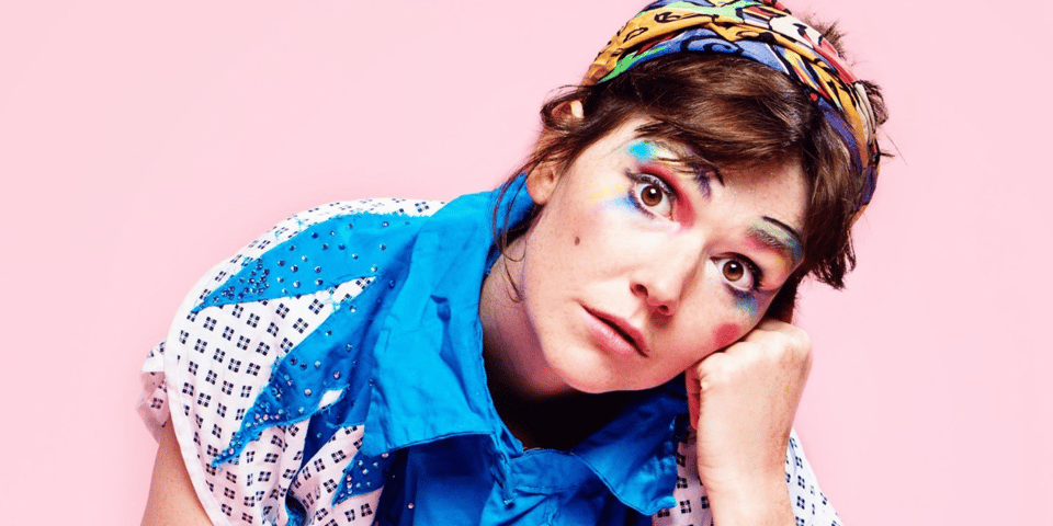 Elf Lyons, who looks like a pixie with rainbow make-up, in front of a pastel pink background.