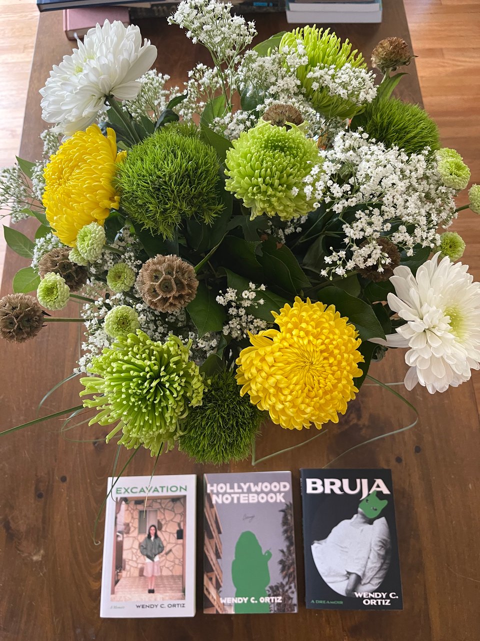 A photo of a wood table with three books on it and a bouquet of beautiful green and yellow and white flowers. The books are Excavation, Hollywood Notebook, and Bruja. They are positioned in a line underneath the bouquet, looking from above.