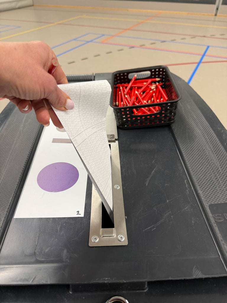 A woman puts her ballot into a receptacle which is a large wheeled plastic garbage container with a metal slit in the top that can be locked